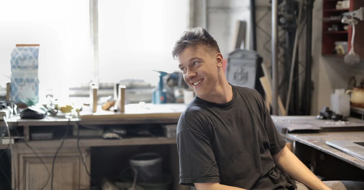 Un jeune artisan souriant assis dans un atelier rempli d'outils et d'équipements.
