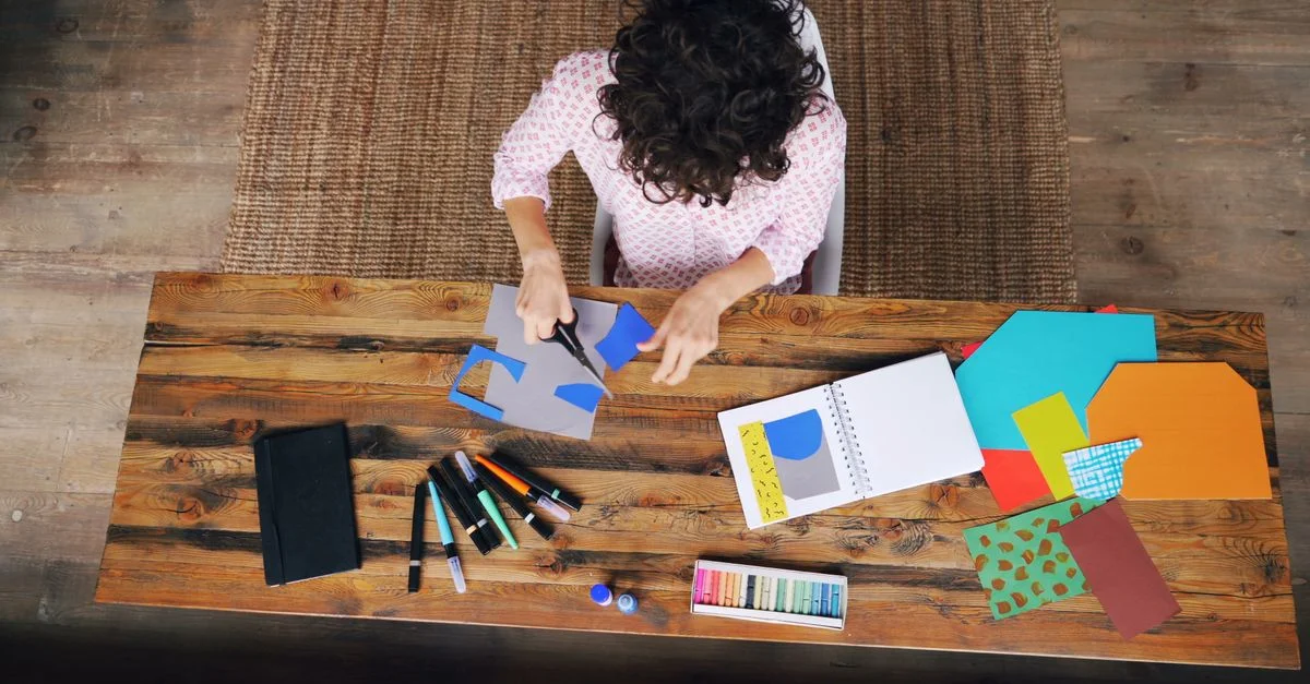 Une personne bricolant avec du papier et des ciseaux, vue de dessus sur une table en bois.