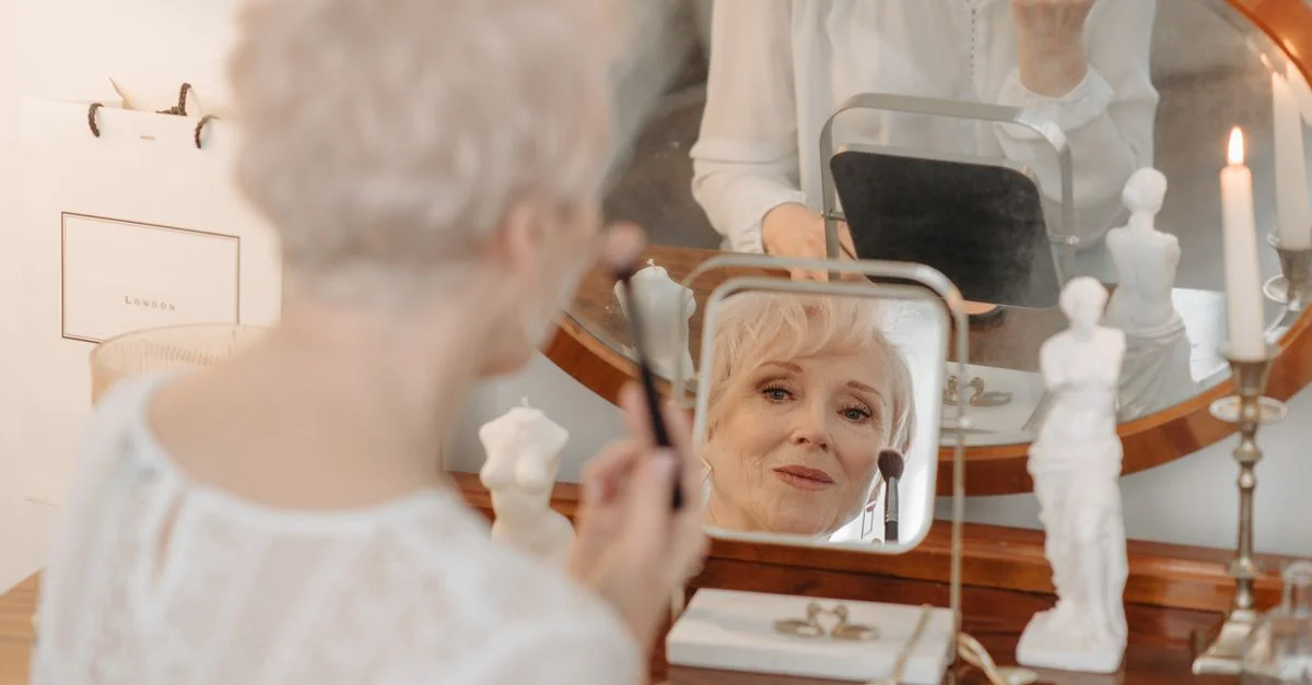 Une femme senior applique du maquillage en se regardant dans un miroir de coiffeuse vintage, créant une atmosphère réfléchissante et sereine.