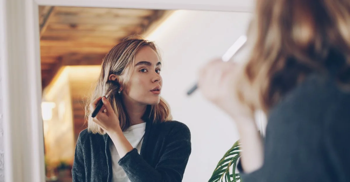 Jeune femme appliquant soigneusement du maquillage avec un pinceau devant un miroir à l'intérieur.