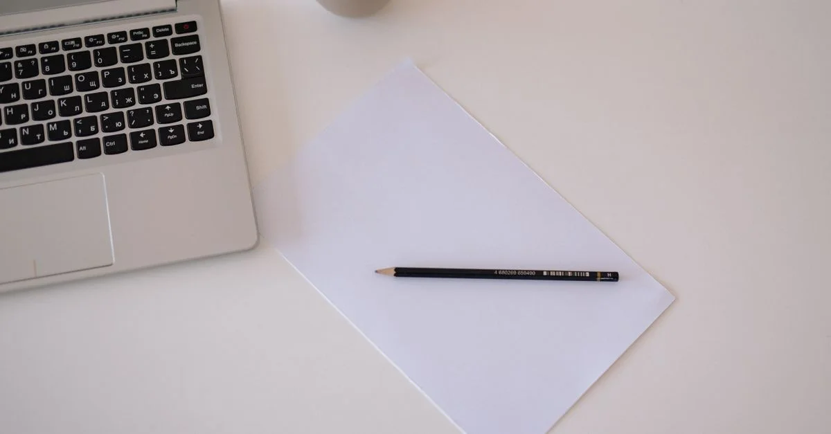 Vue de dessus d'un espace de travail minimaliste avec un ordinateur portable, du papier et un crayon sur une table blanche.