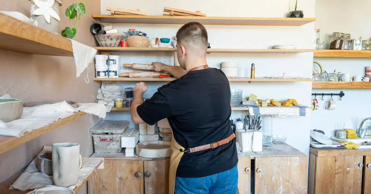 Un artiste céramiste en tablier organise des étagères dans un atelier de poterie accueillant.