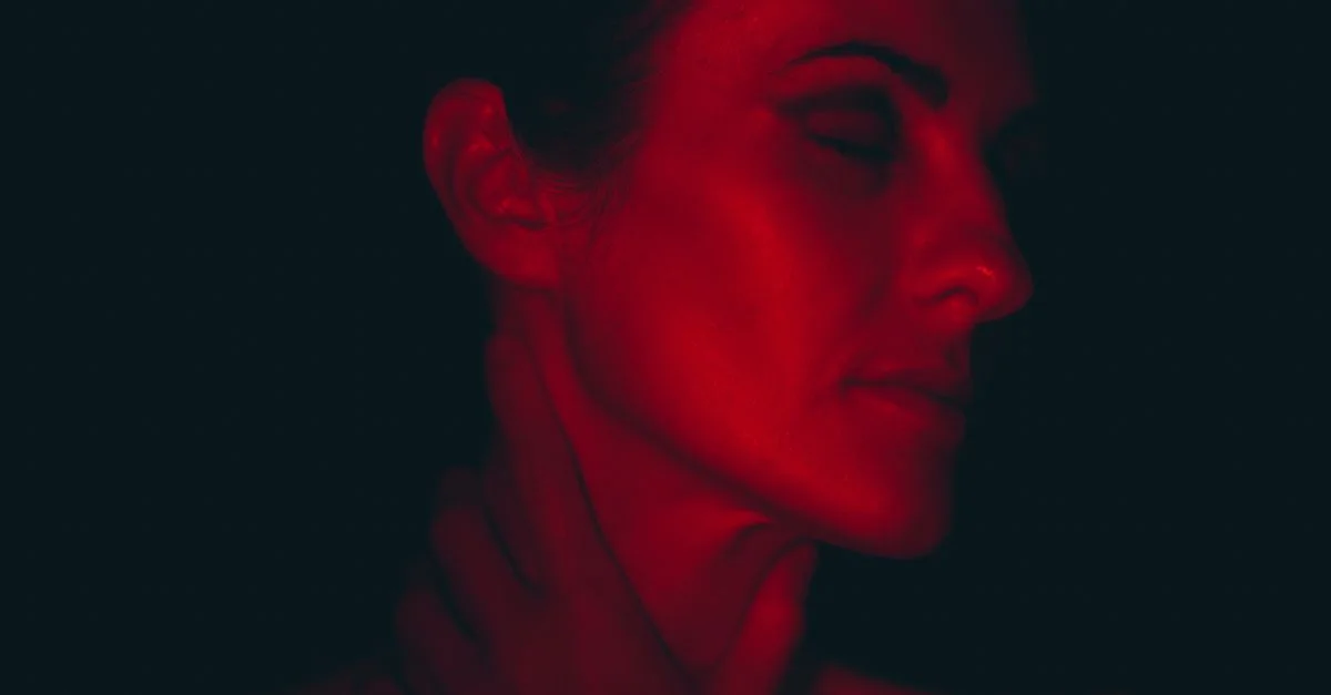 Portrait en gros plan d'une femme les yeux fermés, illuminée par une lumière rouge, créant une atmosphère sombre et artistique.